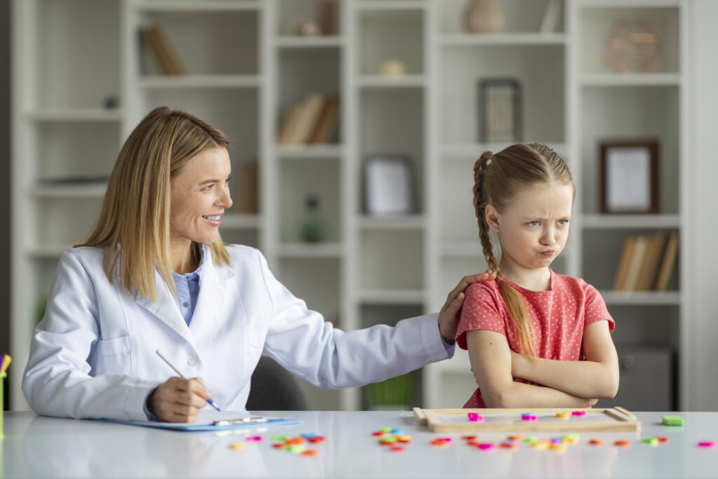 Behavior Therapy For Kids. Smiling Psychotherapist Woman Working With Grumpy Little Girl At Meeting In Office, Offended Female Child Standing With Folded Arms, Ignoring Therapist Lady