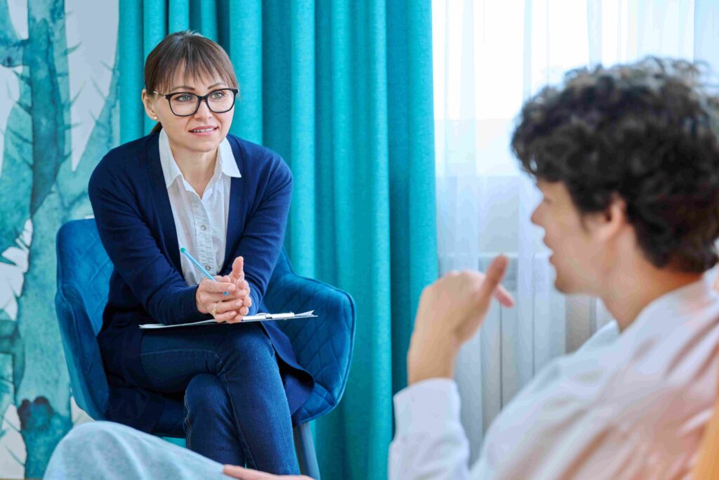 Middle aged confident friendly female psychologist talking to young male, individual therapy session, focus on therapist counselor social worker. Mental health support psychology psychotherapy concept